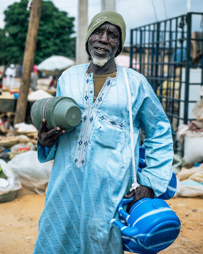 services-04 An elderly man dressed in traditional attire holding household items at an outdoor market, smiling warmly.