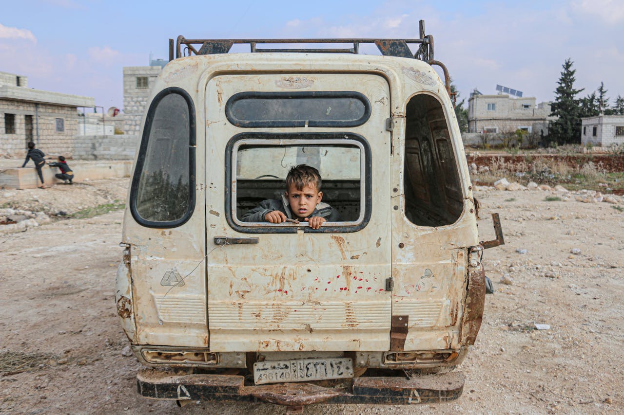 services-01 A young boy peers from an abandoned van in the desolate streets of Idlib, Syria, amidst war devastation.